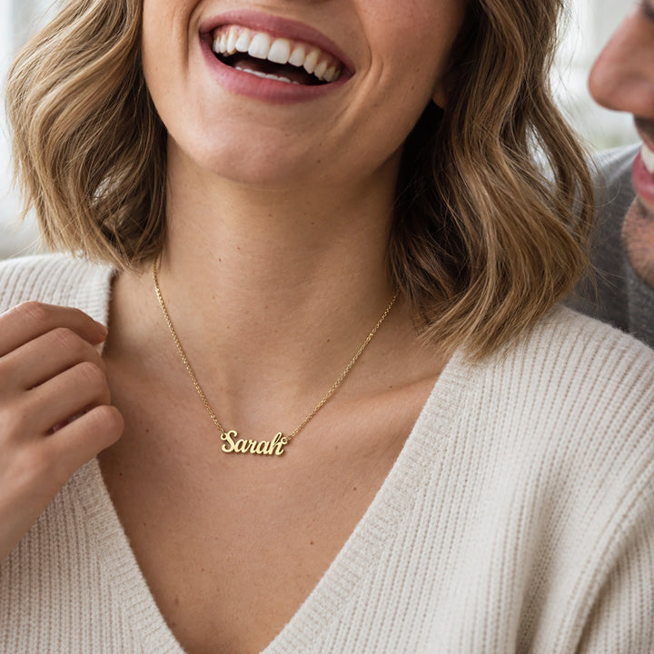 Smiling woman in a cream sweater wearing the gold Name necklace