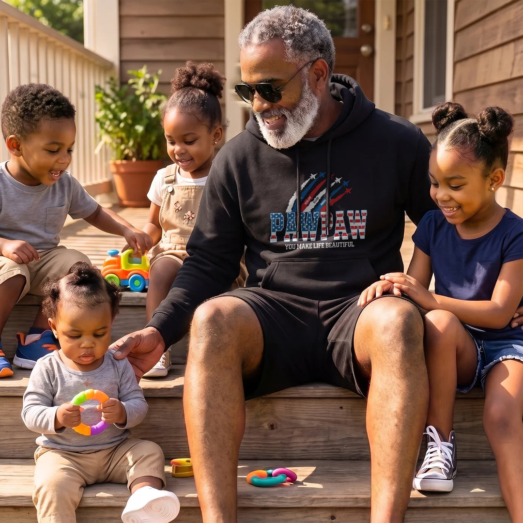 Grandpa in a black PAWPAW American Legend hoodie sits on porch steps with grandchildren
